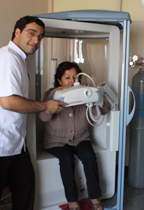 An older woman is seated in a full-sized lung measurement machine, a man stands next to her and helps adjustment the equipment