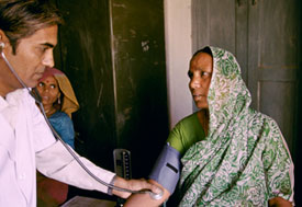 Male medical worker uses stethoscope and arm band to take blood pressure of woman wearing head scarf, another woman in background