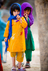 Two young Pakistani girls stand together looking at camera wearing brightly colored outfits and headscarves