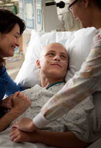 Young man in hospital bed wearing gown, with tubes in nose and no hair, female medical workers stands on each side