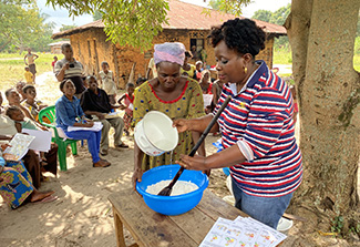 A woman from Kahemba, dressed in a yellow dress, holds a bowl of cassava flour as Dr. Nicole  Mashukano, dressed in a striped pullover and jeans,  pours water into it and stirs. Other villagers, sitting in the background, watch the demonstration.
