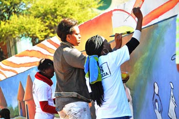 First lady Michelle Obama paints an outdoor mural on a wall between two young African women, sun shines brightly
