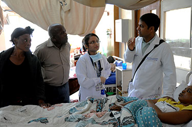 Dr. Omar Siddiqi stands next to bedside of patient, speaks with patient's family and another medical worker.