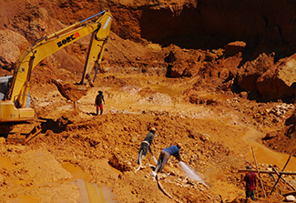 Four workers in large, muddy, open-pit mine. Two spray water next to backhoe, lots of muddy runoff.