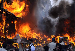 Bright orange flames surrounded by dark black smoke in a city street, buildings in background, people in foreground