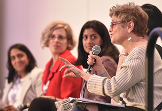 Dr. Michele Barry speaks into a microphone while seated on a dais with four women during a panel discussion at a conference