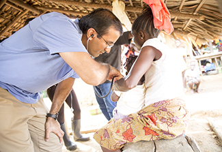The photo shows Dr. Raj Panjabi, wearing a blue shirt and tan slacks, attending to a patient, a tween-aged girl in a white blouse and beige patterned skirt.  The photo was taken in the girl’s home and the background shows a thatched roof. 