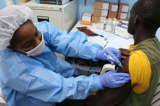 In a clinic, patient receives a shot in his arm from a medical worker in gloves and a protective mask, head covering and gown.