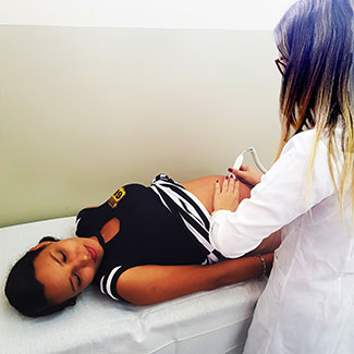 Pregnant teenage girl lies on her back in an exam room while a medical worker with her back to the camera examines her belly