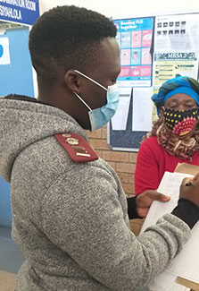 Researcher wearing a mask takes notes on a clipboard while interviewing a masked subject in a clinic.