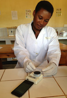 Woman wearing white lab coat and gloves adjust the mChip prototype while it is connected to a smartphone on a counter in a lab