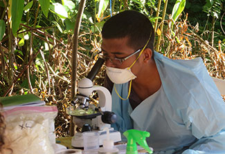 A researcher in protective mask and clothing peers into a microscope in an outdoor lab with trees in the background.