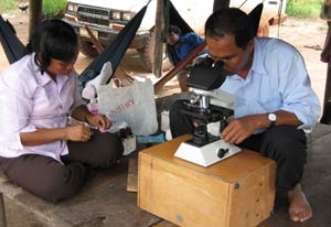 Man squatting looks through microscope, woman seated prepares samples, both on wooden platform with open air sides
