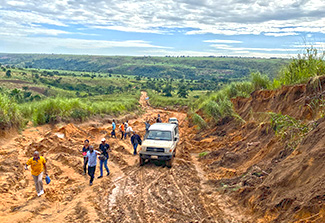 The photo shows a van driving on a muddy, rutted road alongside walking villagers on the way to Kahemba.