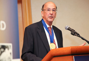 Roger Glass speaks into a microphone at a podium wearing the Sabin Gold Medal around his neck