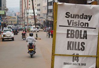 Sign shows newspaper headline from 2012 Ebola Kills 14 with busy city street in the background.