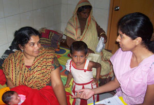Woman seated holding newborn, young child stands next to her, researcher with papers in lap speaks to mother, woman in backgroun