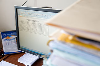 Stacks of paper on desk next to computer monitor showing data