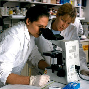 Two healthcare workers in lab wearing white coats, one looks through microscope, taking notes, another observes