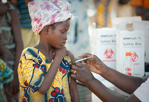 Young girl in outdoor clinic holds up sleeve to receive shot in the upper arm, healthcare worker off camera