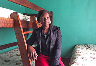 The photograph shows a teenaged girl dressed in black and red clothes. She is leaning against a bunk bed ladder inside a women’s shelter in Zimbabwe where she hopes to receive aid and counseling.