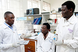 Three male medical workers in white coats wearing gloves, one explains to the other two