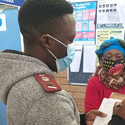 Researcher wearing a mask takes notes on a clipboard while interviewing a masked subject in a clinic.