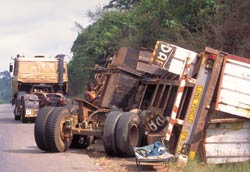 Large truck rolled over on side on the side of a paved road, wheels and axels detached, truck severely damaged