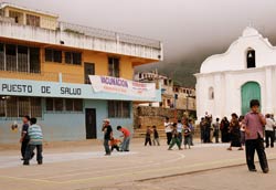 Children play on paved courtyard, buildings in background, sign reads puesto de salud