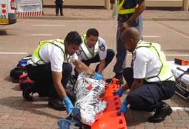 Three male emergency response workers in uniforms carefully roll wrapped, adult-sized body from brick pavement onto stretcher