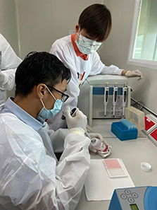 Two people in white lab coats and masks work at a lab bench: one in the foreground wearing glasses handles a sample with gloved hands, while another stands behind operating a piece of laboratory equipment with attached tubes, with pipette tip boxes and other supplies nearby.