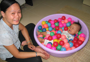 Baby lies in purple bucket full of balls, woman seated cross-legged on floor