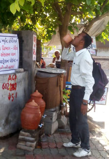 In India, a man drinks from a cup at a public outdoor drinking water station in the shade on a street corner