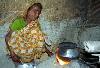 Woman squats next to pot cooking over an open flame, thatched walls in background