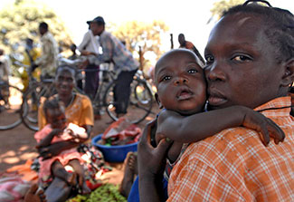 Woman holding baby outdoors looks at camera, another woman and baby and men on bikes in background.