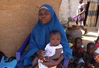 Woman, seated, holds young child on her lap, more children seated on the ground in the background.