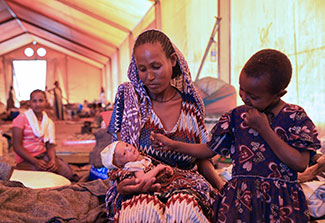 Woman holds a newborn baby, older child seated next to her on a cot, in a large tent clinic, other women in background.