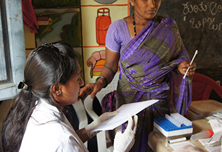 A woman dressed in a purple sari returns her self-collection vaginal swab to a clinic worker, dressed in medical whites, for HPV testing 