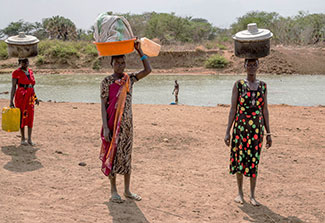 Three women carry large water containers on their heads and in their hands, standing in front of a body of water.