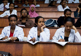 Three female students in white lab coats taking notes in large lecture hall, many other women seated in background