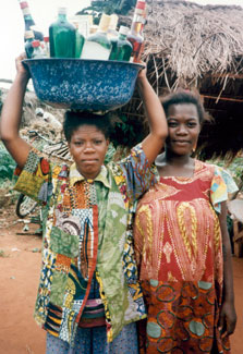 Two African women outdoors, one balances tub full of alcohol bottles on her head