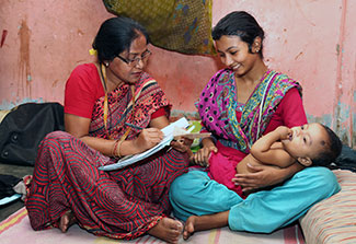 A researcher interviews and takes notes while talking to a woman holding an infant