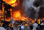 Bright orange flames surrounded by dark black smoke in a city street, buildings in background, people in foreground