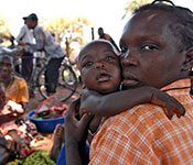 By UN Photo/Tim McKulka. Woman holding baby outdoors looks at camera, another woman and baby and men on bikes in background.