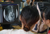 Two women with backs to camera review a medical image on a monitor in a crowded computer lab