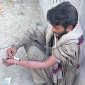 Pakistani man squats on ground against wall while injecting need into forearm