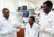 Three male medical workers in white coats wearing gloves, one explains to the other two