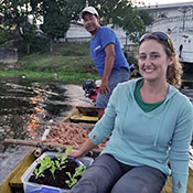 Dr. Leann Andrews seated in small boat on the water next to seedlings in soil.