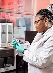 Female researcher working wearing lab coat and gloves examines testing supplies in front of lab equipment in a CAPRISA lab.