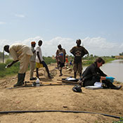 In Senegal, six researchers, some wearing gloves and boots, standing and seated on dry ground next to calm body of water.